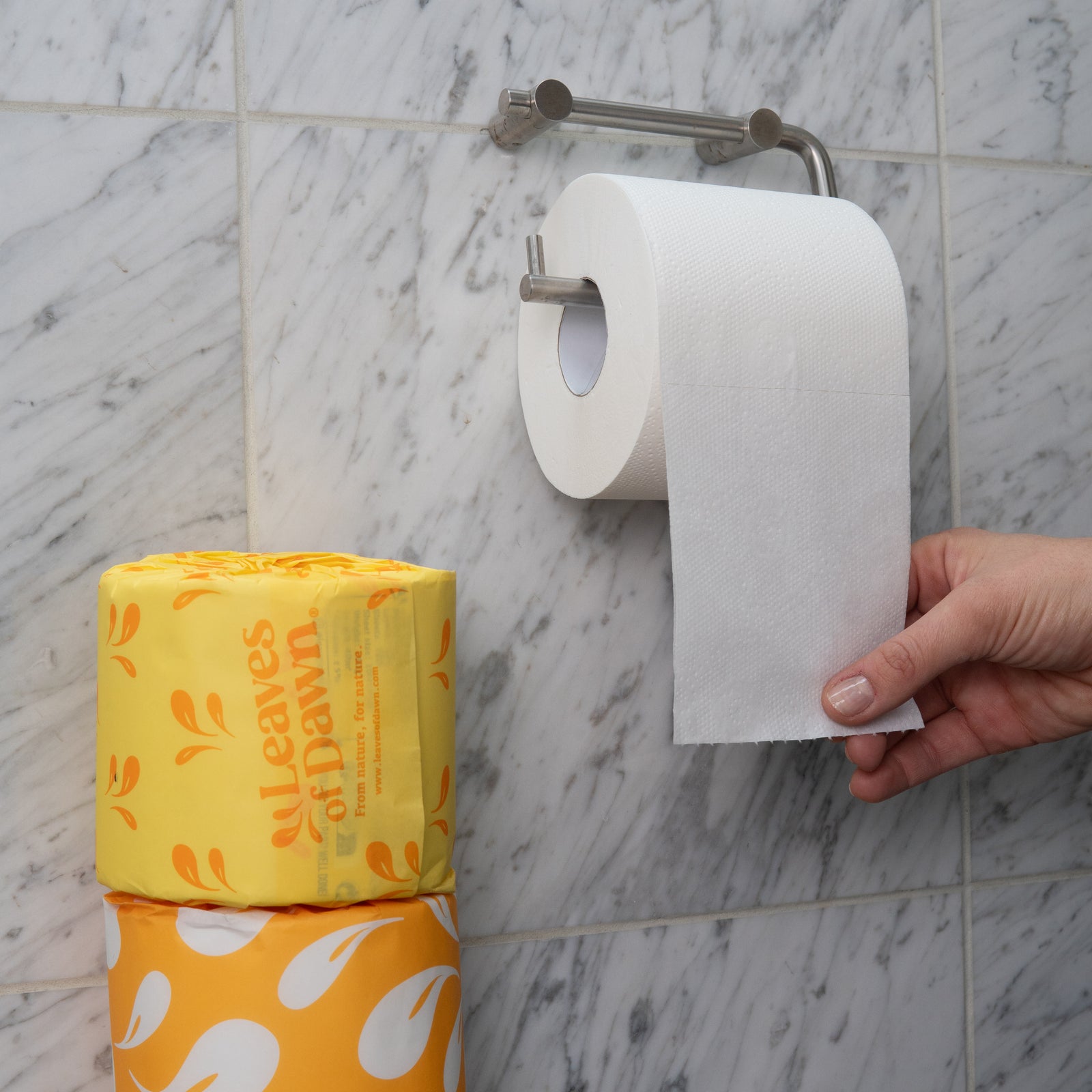 Leaves of Dawn premium 3-ply bamboo toilet paper rolls on a toilet paper holder,  a hand pulling the paper, showing its soft texture and whiteness.

